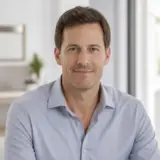 Portrait d’Aurélien Pradeau, cheveux châtains courts, chemise bleu gris, sourire discret, fond de pièce lumineuse et floue