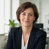 Portrait de Nadia Sauvaget, cheveux bruns courts, veste sombre, léger sourire, fond de bureau clair et flou